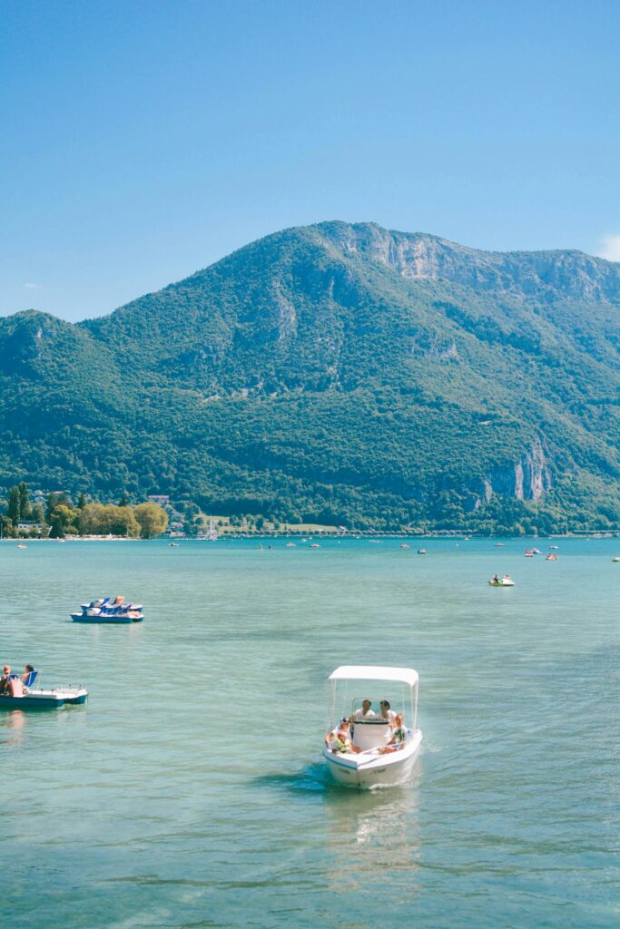 Panoramic view of Lake Annecy in the French Alps, featuring crystal-clear turquoise waters, lush green hills, and distant snow-capped mountains under a bright blue sky.