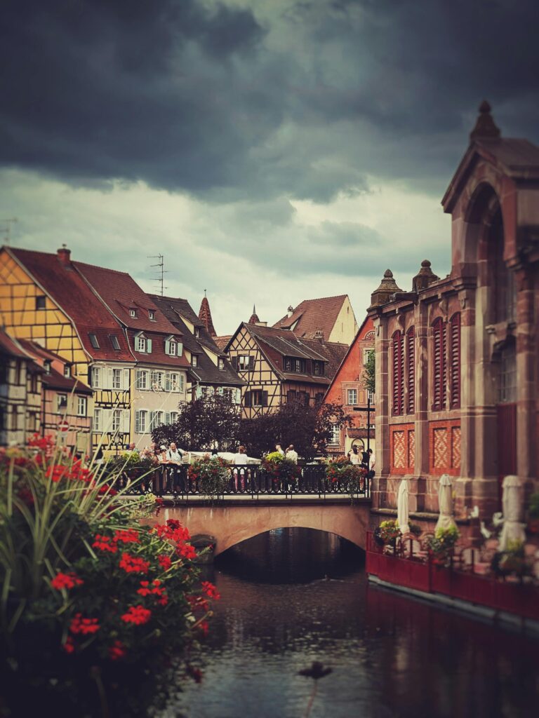 Beautiful view of Colmar, France, with vibrant flowers in the foreground, colorful half-timbered houses, and charming cobblestone streets in a picturesque village setting.